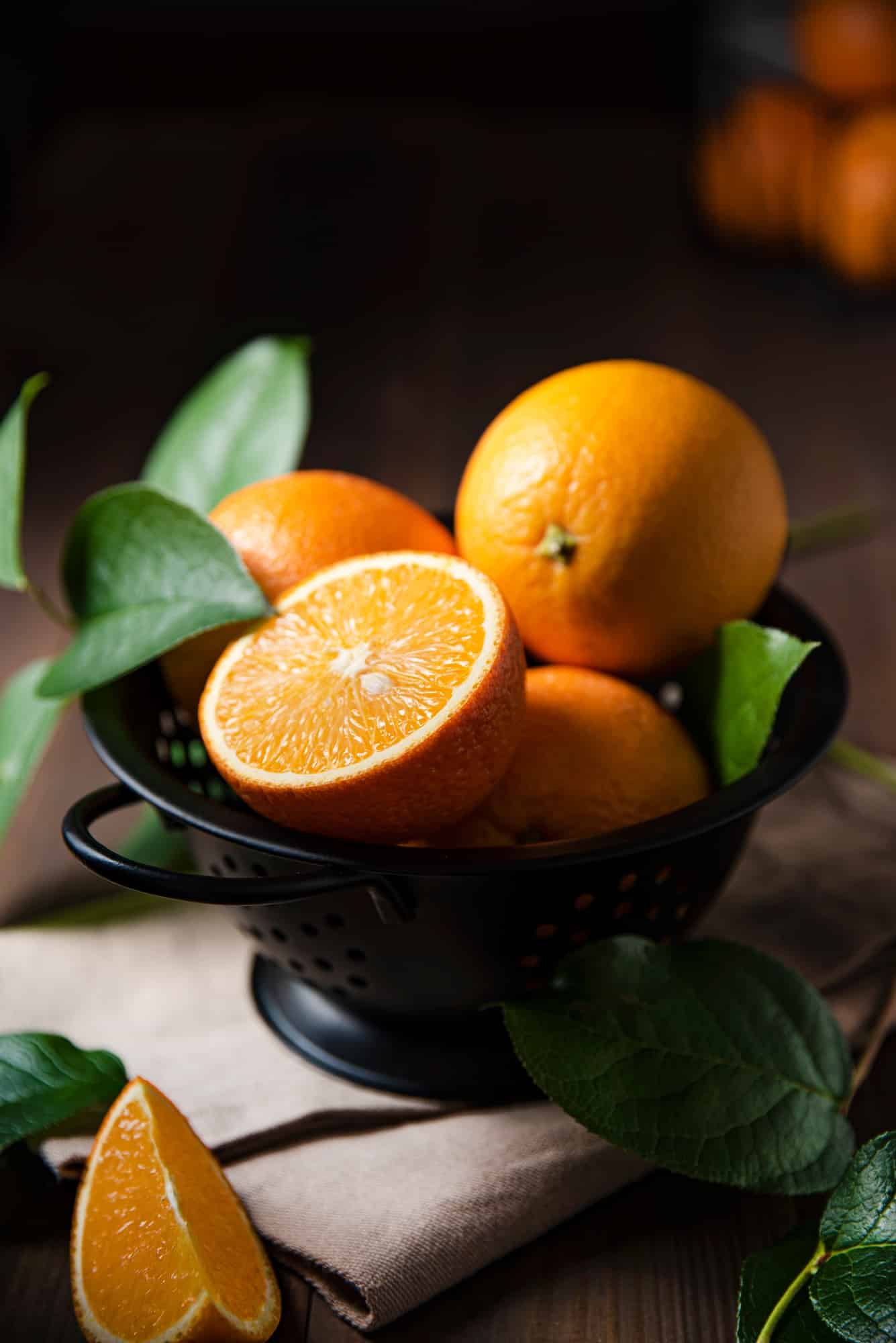 A few juicy oranges in a black bowl with green leaves on a dark wooden background