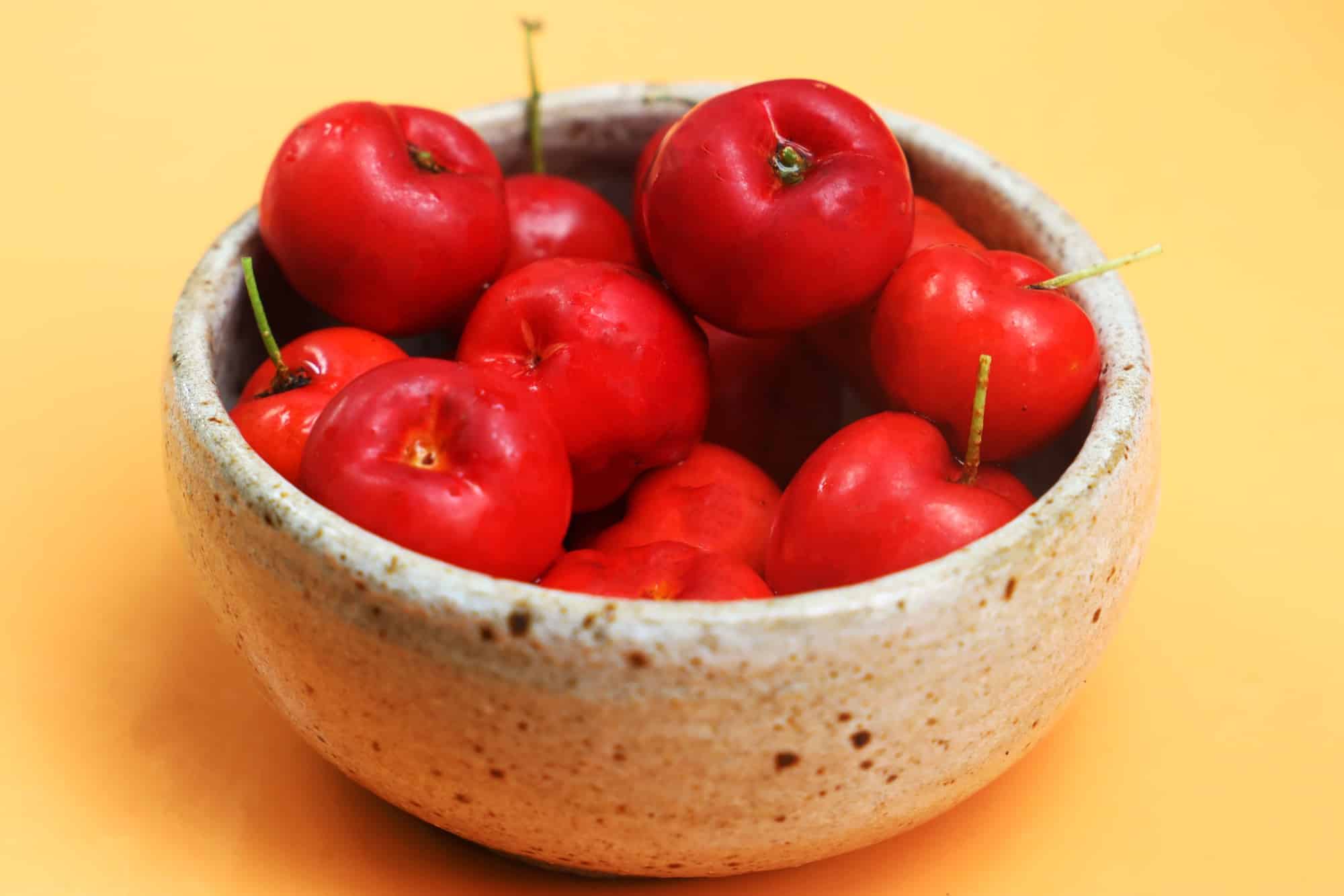 Bowl of fresh acerola cherries on yellow background.